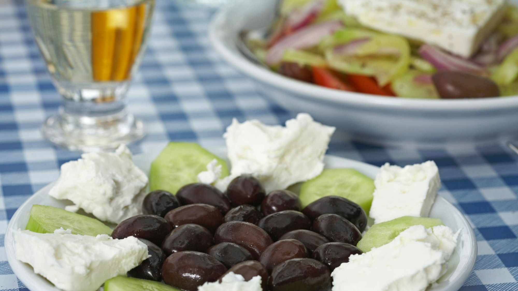 Teller mit Gurken, Oliven und Feta und einem Glas Retsina auf einer blau-weißen Tischdecke