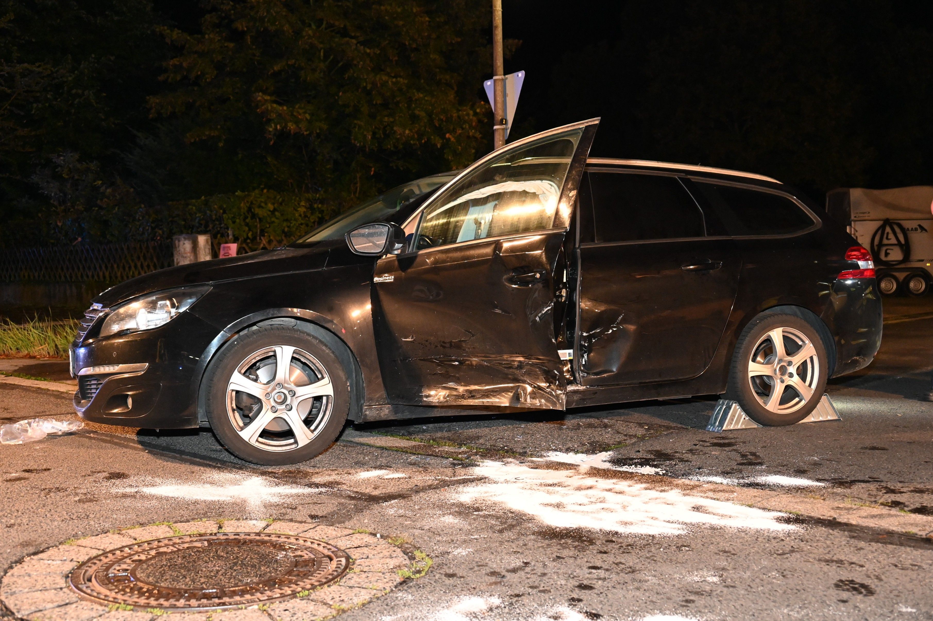 Ein dunkles Auto steht mit eingedellter Seite und offener Fahrertür an der Unfallstelle.