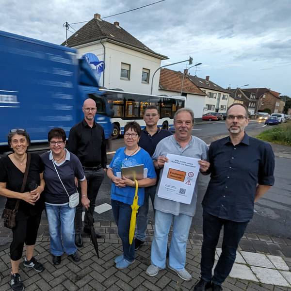 Eine Gruppe von Männern und Frauen mit Protestplakat an der Bonnstraße, im Hintergrund fahren Lkw, Bus und Autos vorbei.