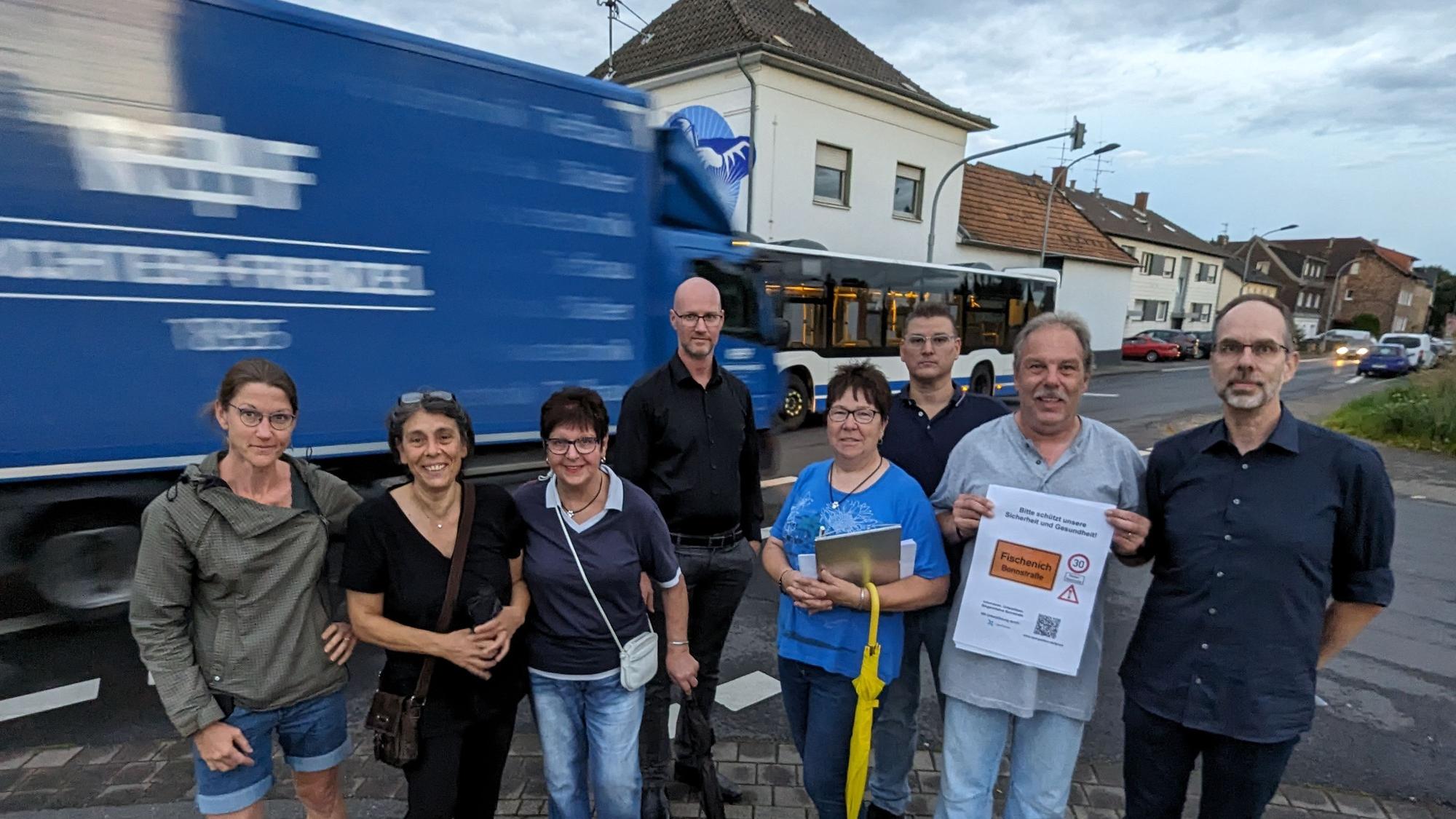 Eine Gruppe von Männern und Frauen mit Protestplakat an der Bonnstraße, im Hintergrund fahren Lkw, Bus und Autos vorbei.