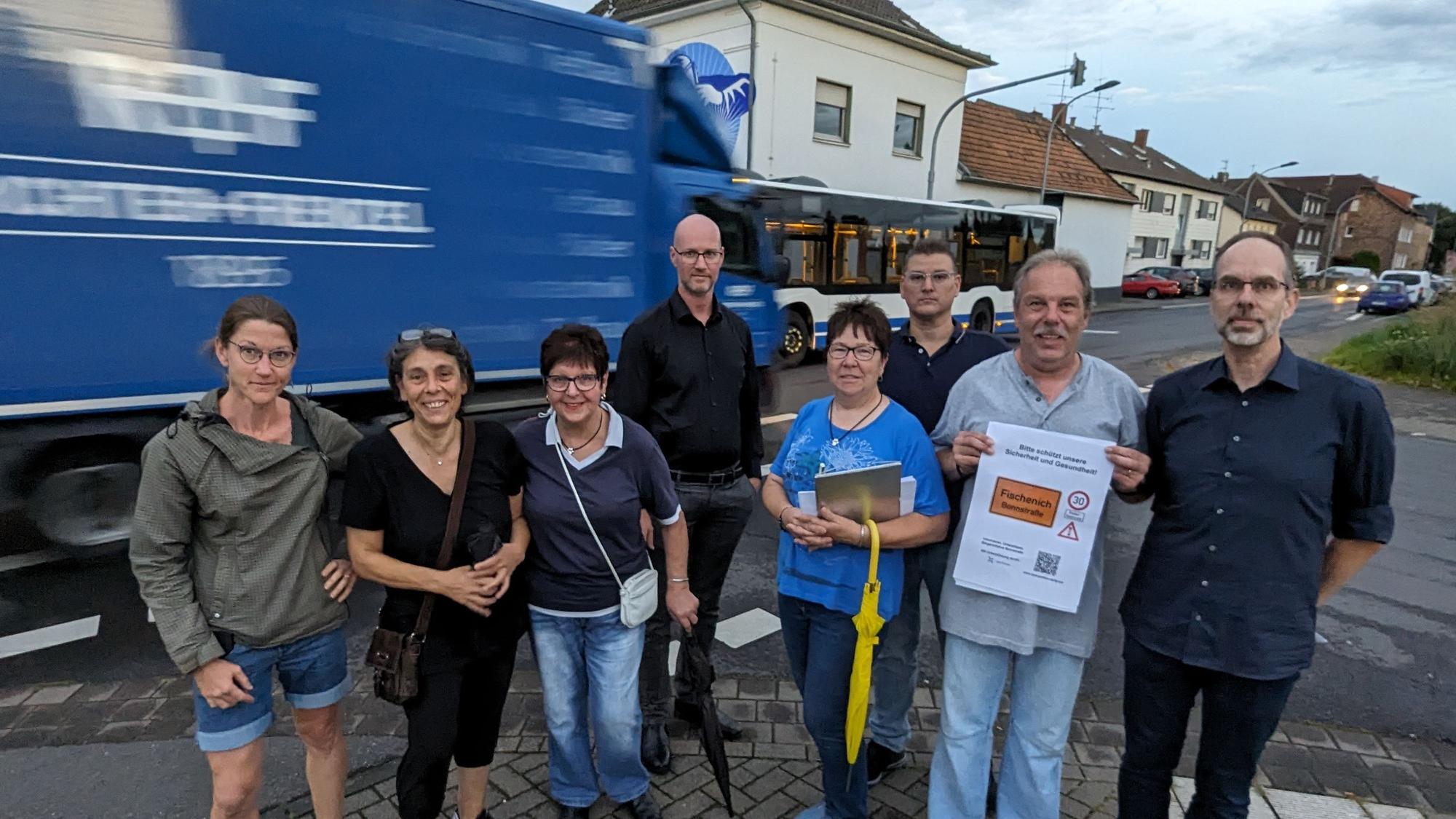 Eine Gruppe von Männern und Frauen steht an einer Straße, im Hintergrund ein Lkw und ein Bus.