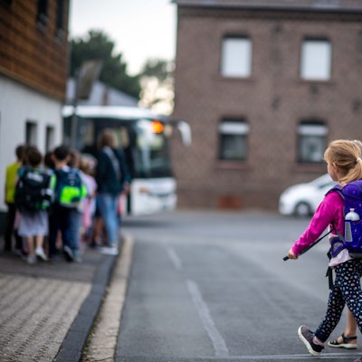 Das Bild zwei Grundschülerinnen, wie sie eine Straße überqueren. Im Hintergrund geht eine weitere Gruppe in Richtung Bus.