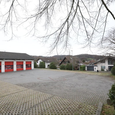 Ein weißes Haus mit vier roten Rolltoren; Fahrzeuge der Feuerwehr sind dahinter geparkt. Auf dem Dach steht eine Feuerwehrsirene.