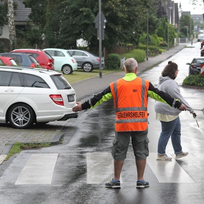 Ein Schülerlotse in oranger Warnweste steht an einem Zebrastreifen, Kinder überqueren ihn, ein Auto parkt gerade aus.