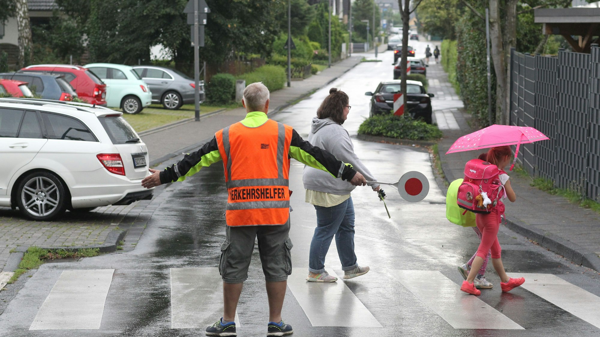 Ein Schülerlotse in oranger Warnweste steht an einem Zebrastreifen, Kinder überqueren ihn, ein Auto parkt gerade aus.