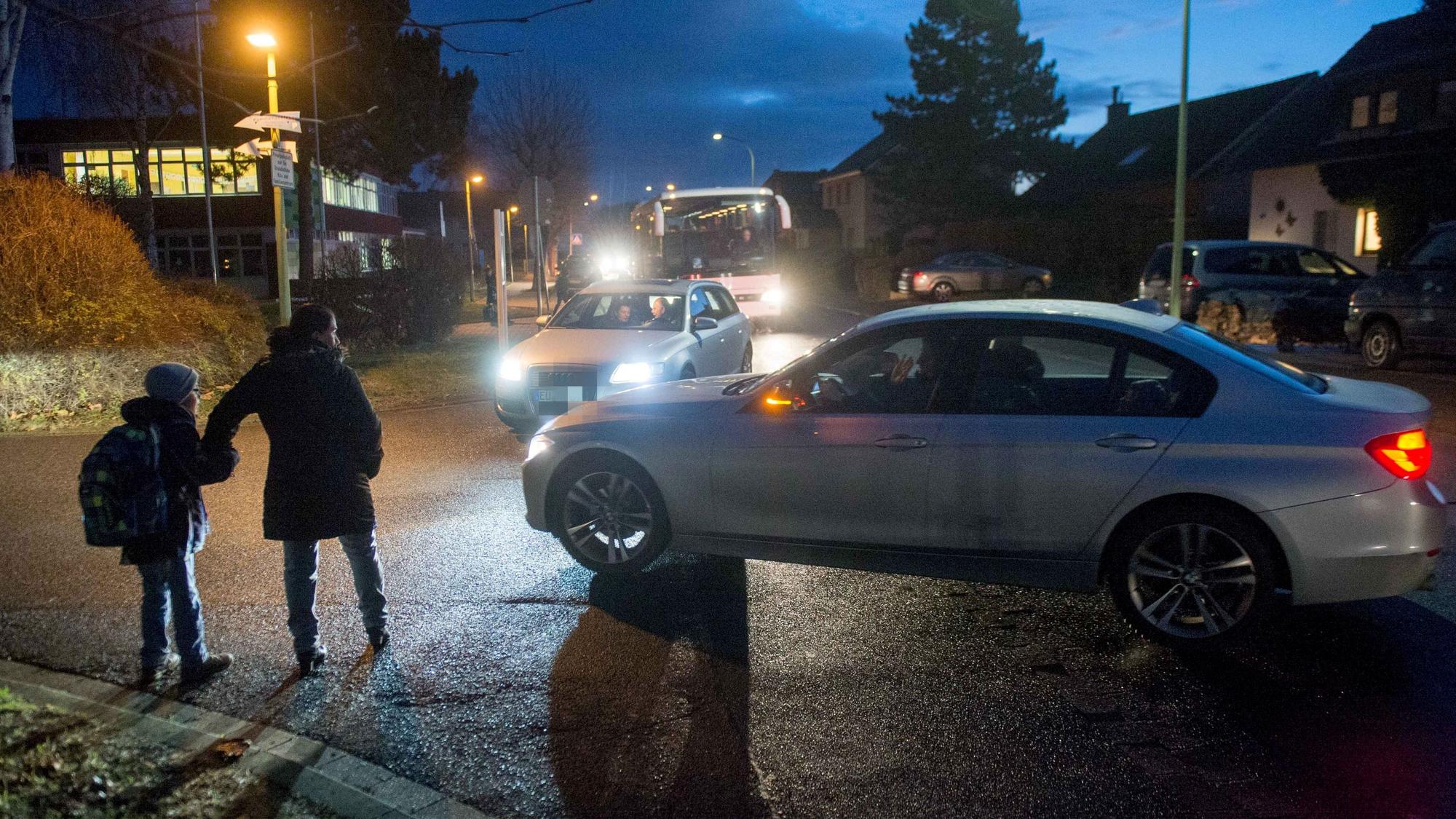 Das Bild zeigt eine gefährliche Situation vor der Schule. Zwei Autos wollen in eine Straße abbiegen, übersehen dabei aber ein Kind mit seiner Mutter.