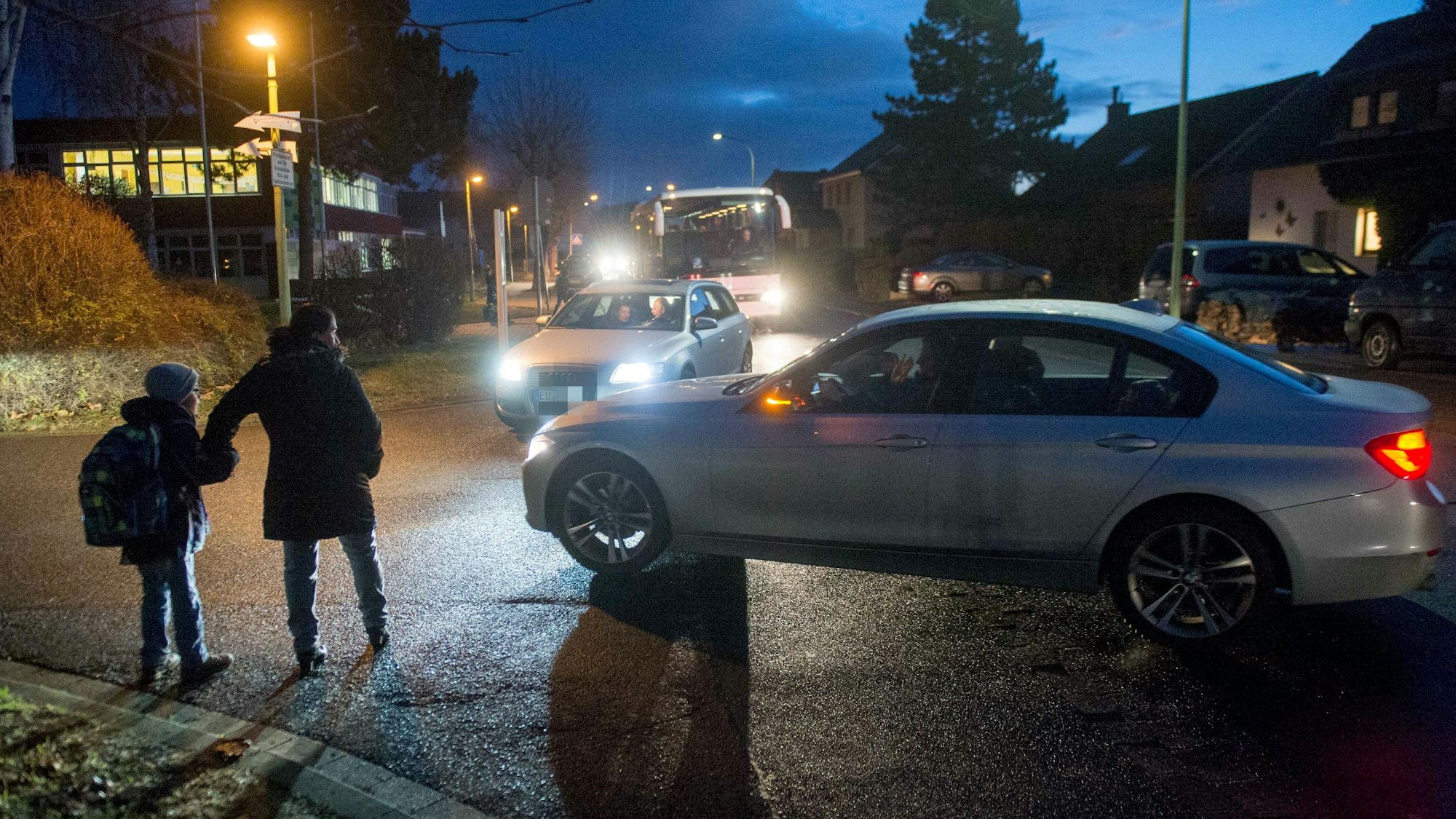 Das Bild zeigt eine gefährliche Situation vor der Schule. Zwei Autos wollen in eine Straße abbiegen, übersehen dabei aber ein Kind mit seiner Mutter.
