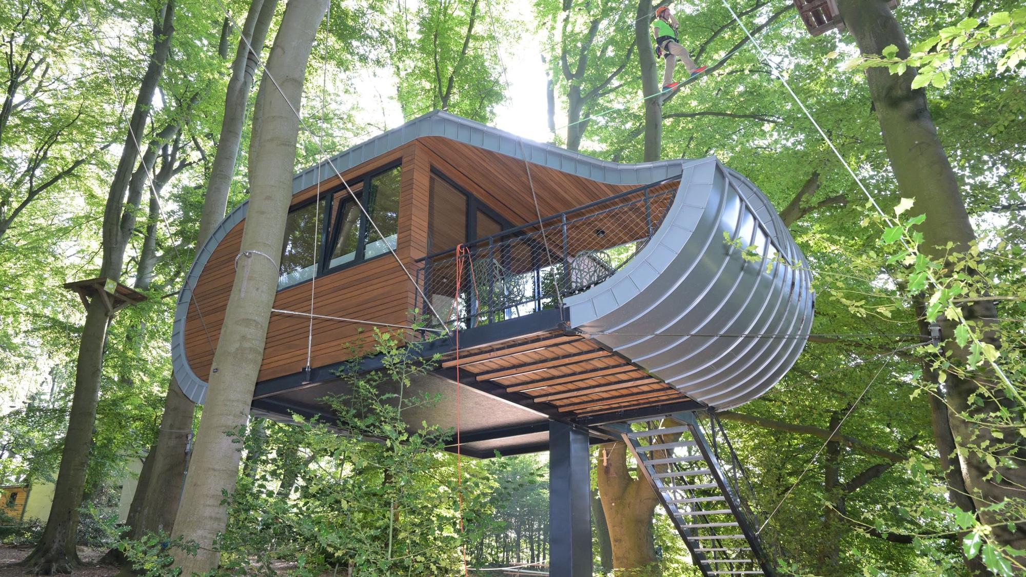 Ein schlittenartiges Baumhaus hängt mit Stahlseilen befestigt an großen Buchen.