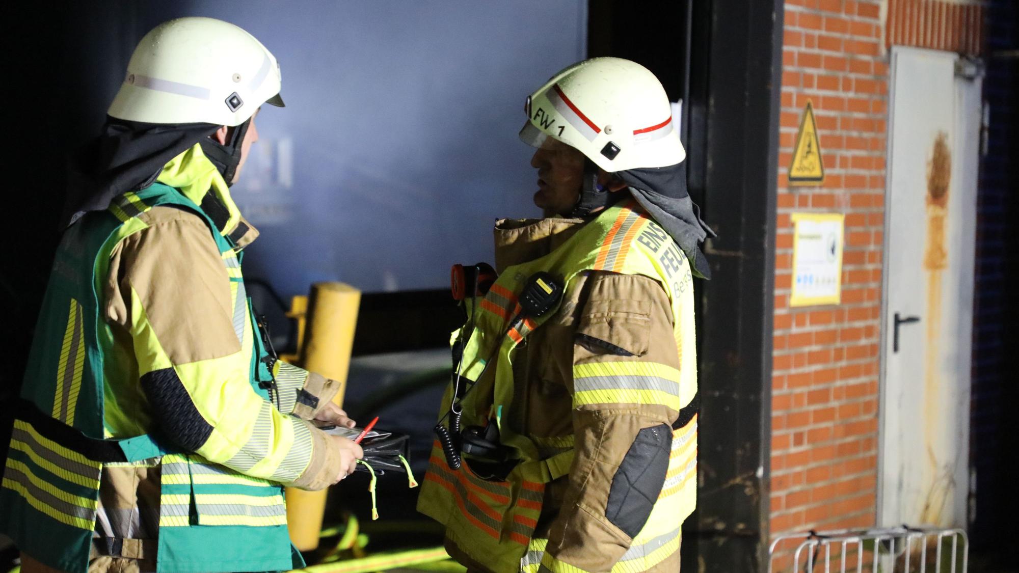 Zwei Feuerwehrleute stehen vor einer verrauchten Lagerhalle.