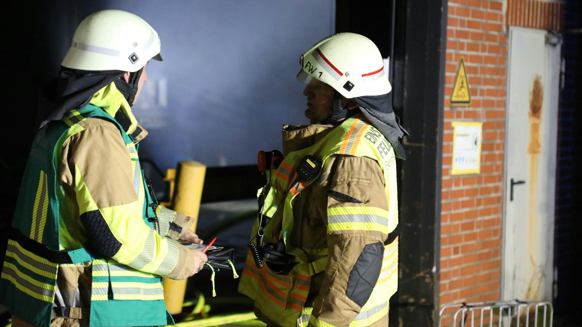 Zwei Feuerwehrleute stehen vor einer verrauchten Lagerhalle.