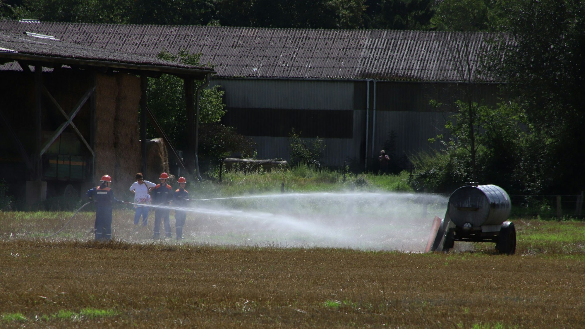 Kinder in blauen Overalls und mit orangefarbenen Helmen richten Löschschläuche auf einen Güllewagen.