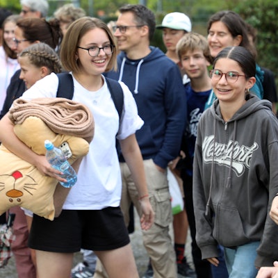 Schülerinnen und Schüler, Lehrerinnen und Lehrer und Eltern und andere Menschen stehen mit Fahnen von Ukraine, Deutschland, Butscha und Bergisch Gladbach auf dem Schulhof der Integrierten Gesamtschule Paffrath und jubeln.