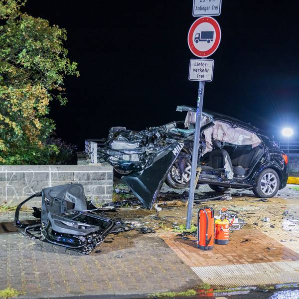 Ein völlig verformtes Autowrack häng an einer etwa 90 Zentimeter hohen Mauer halb in er Luft, Autoteile liegen rundum, Strahler der Einsatzkräfte beleuchten den Unfallort.