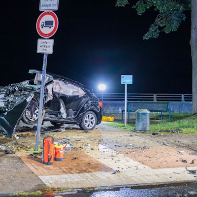 Ein völlig verformtes Autowrack häng an einer etwa 90 Zentimeter hohen Mauer halb in er Luft, Autoteile liegen rundum, Strahler der Einsatzkräfte beleuchten den Unfallort.