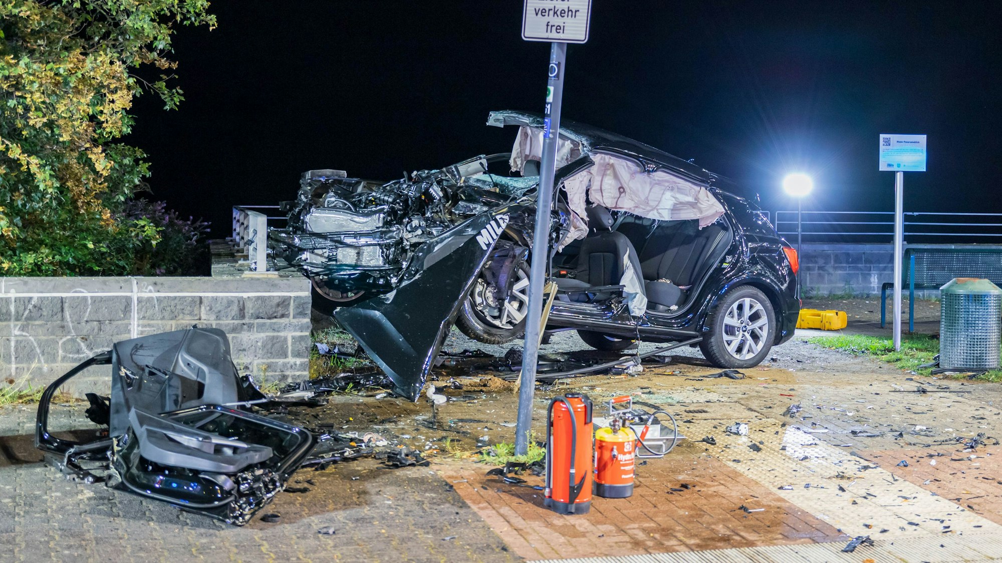 Ein völlig verformtes Autowrack häng an einer etwa 90 Zentimeter hohen Mauer halb in er Luft, Autoteile liegen rundum, Strahler der Einsatzkräfte beleuchten den Unfallort.
