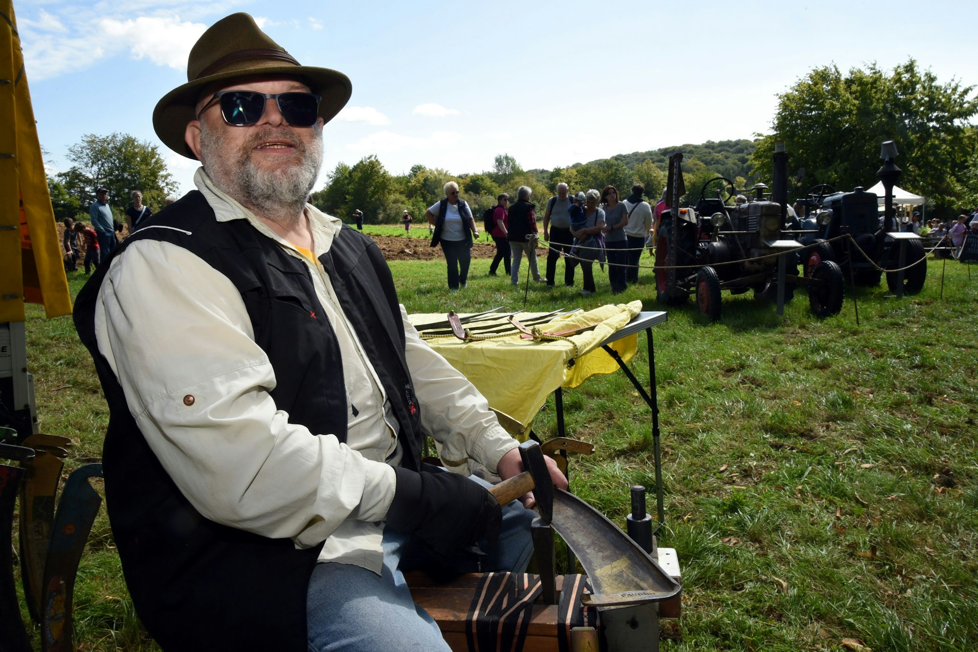 Ein Mann dengelt eine Sense, im Hintergrund Zuschauerinnen und Zuschauer.