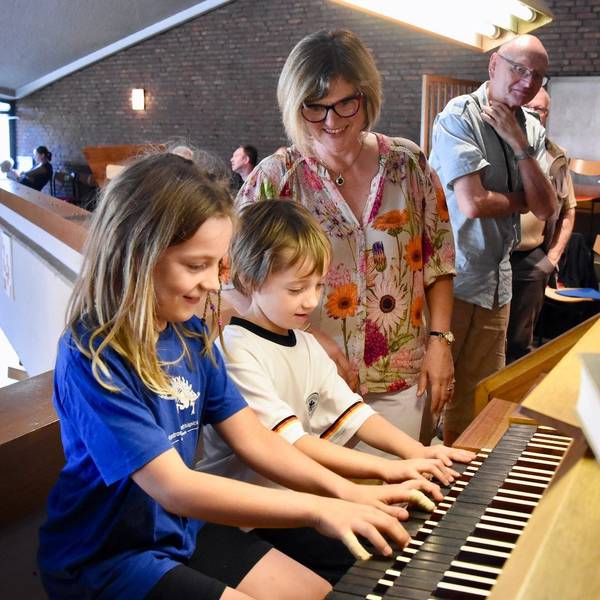 Ein Junge und ein Mädchen spielen auf einer Orgel. Eine Frau in bunter Bluse schaut, im Hintergrund steht ein weiterer Zuschauer.