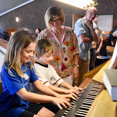Ein Junge und ein Mädchen spielen auf einer Orgel. Eine Frau in bunter Bluse schaut, im Hintergrund steht ein weiterer Zuschauer.