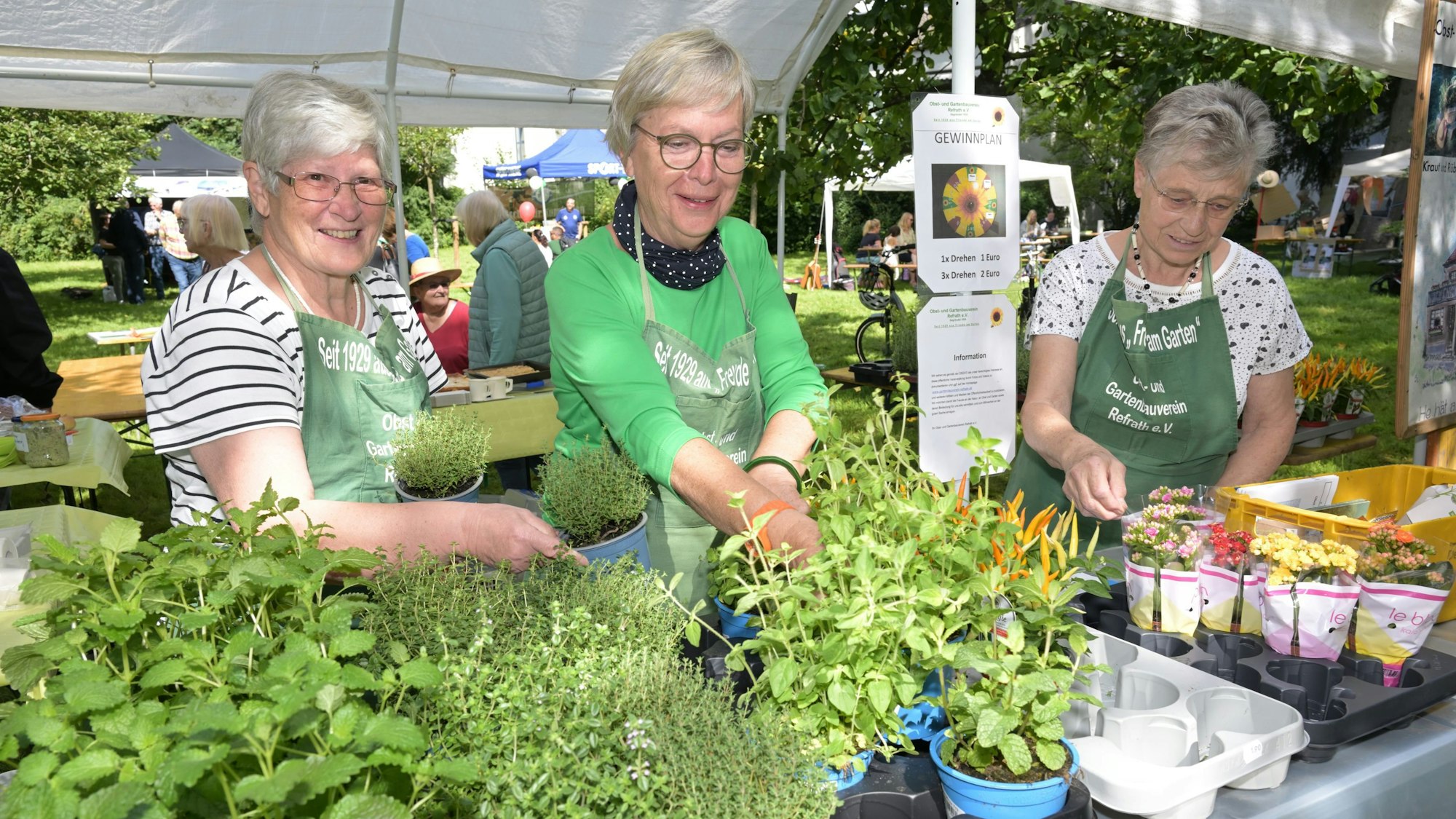Drei Frauen mit Gärtnerschürze an einem Stand mit Kräutern und Blumen.