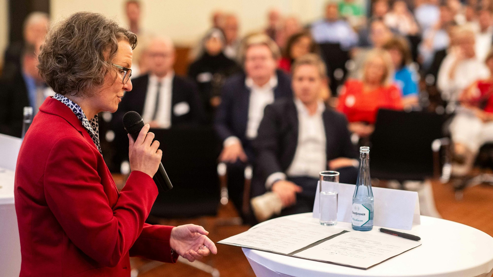 Das Bild zeigt Ministerin Ina Scharrenbach mit einem Mikrofon in der Hand, wie sie zum Publikum spricht.