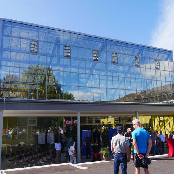 Vor einem großen gläsernen Gebäude stehen Besucher. In den Glasscheiben spiegeln sich blauer Himmel, Bäume und Wolken.