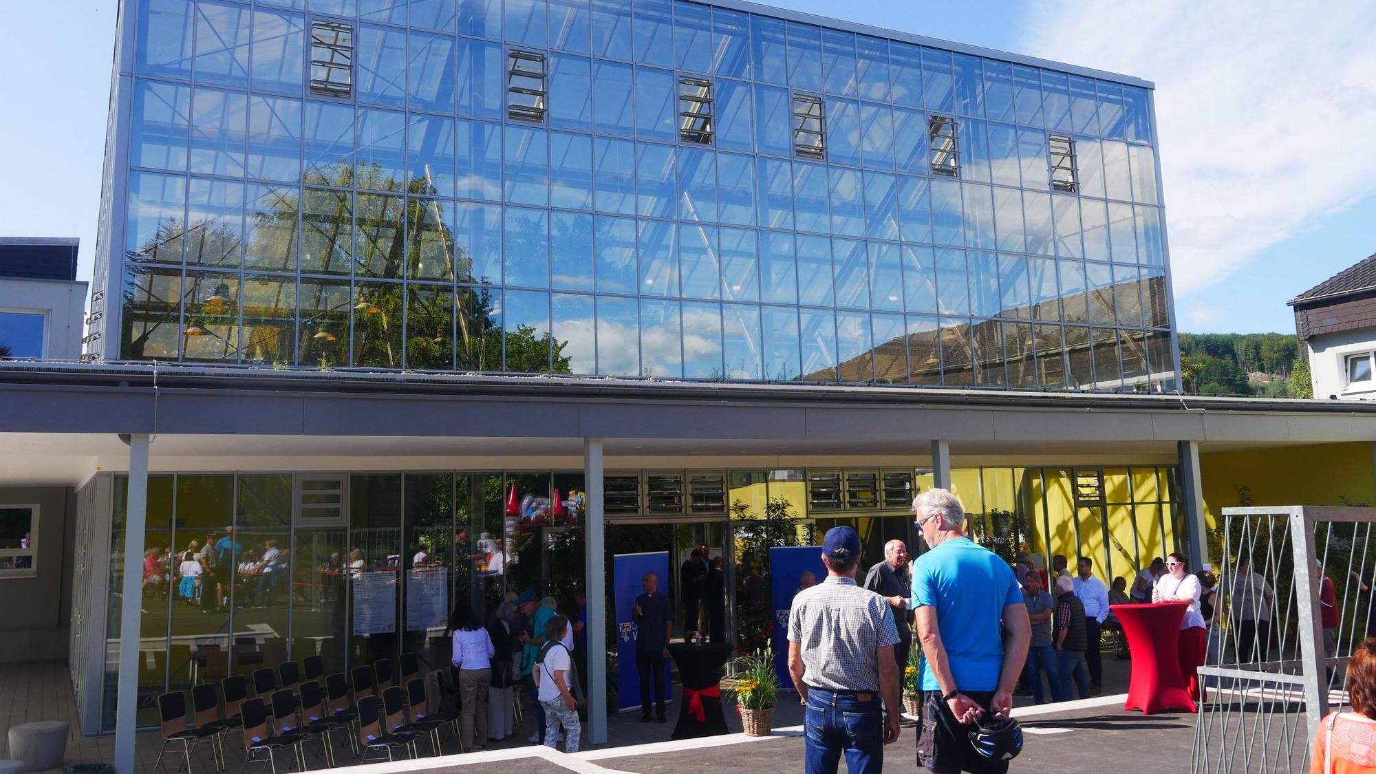 Vor einem großen gläsernen Gebäude stehen Besucher. In den Glasscheiben spiegeln sich blauer Himmel, Bäume und Wolken.