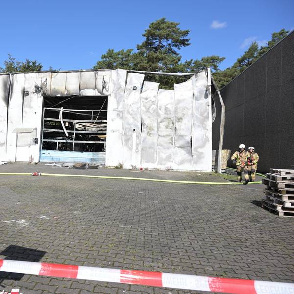 Absperrband prangt vor einer ausgebrannten Halle, Feuerwehrleute gehen auf einem Kontrollgang um die Halle.