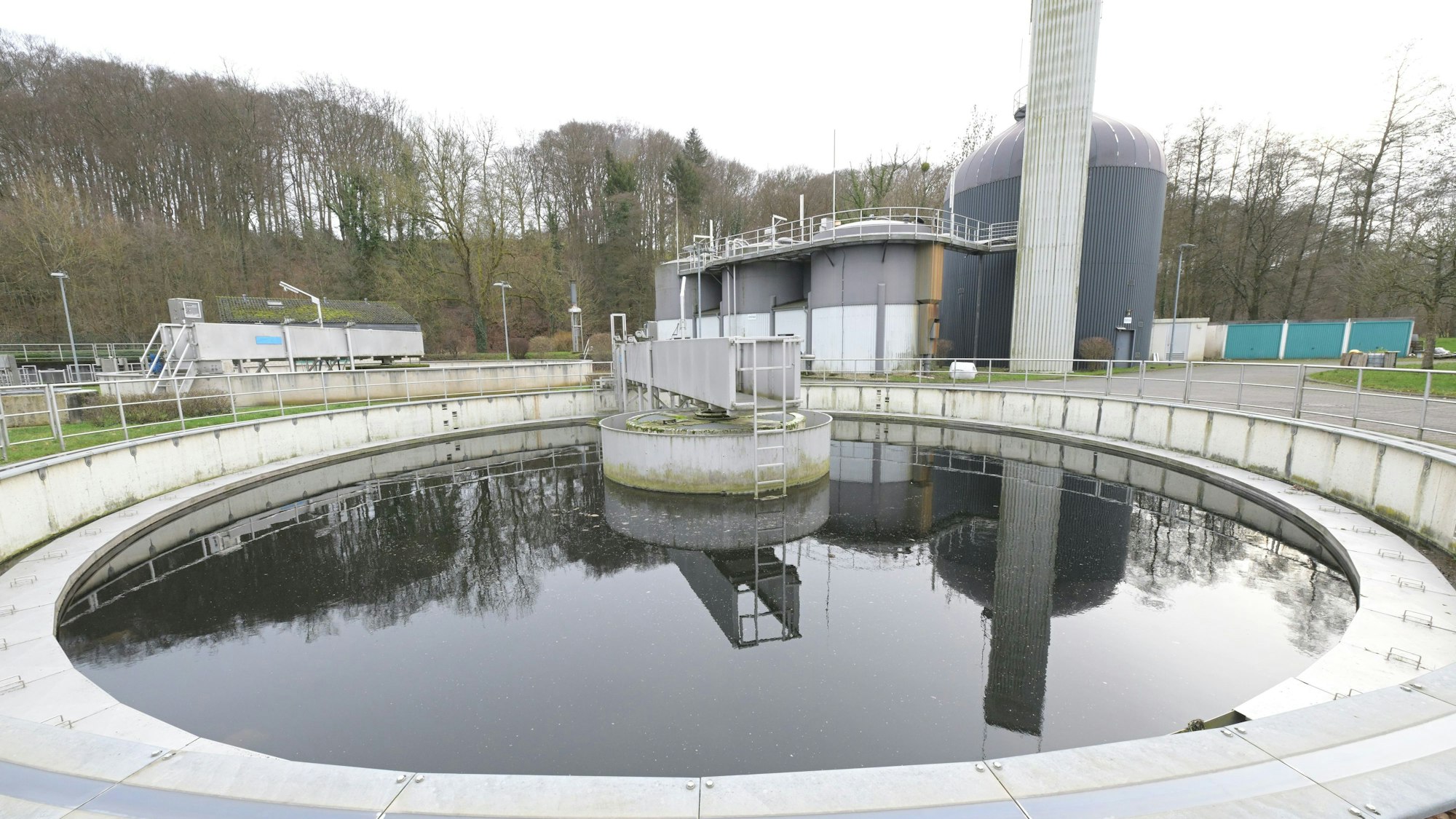 Ein kreisrundes Becken einer Kläranlage, dahinter mehrerer kleine Faultürme.