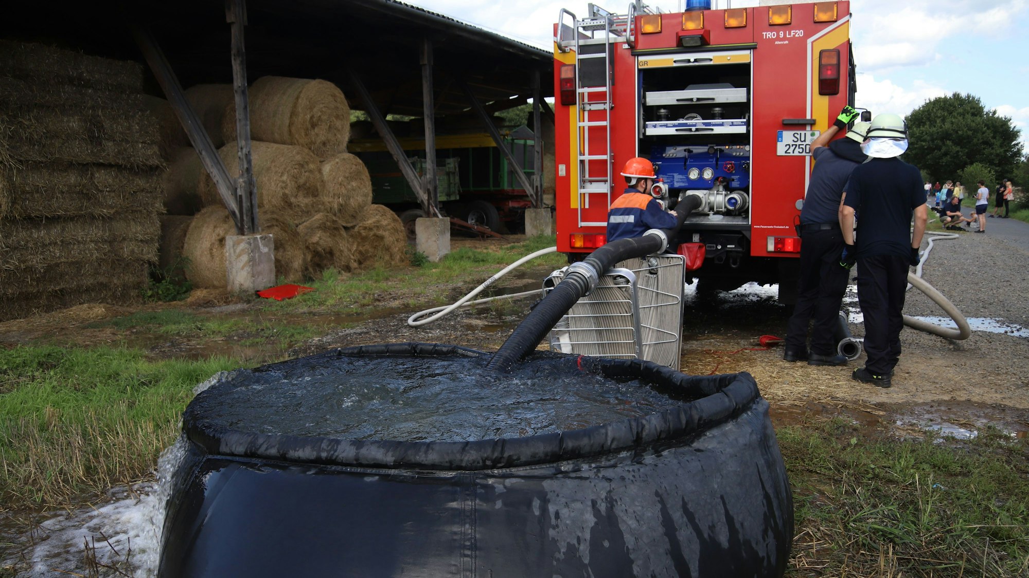 Ein Feuerwehrauto ist über einen Schlauch mit einem schwarzen Faltbecken für Wasser verbunden. Links sind Strohballen unter einem Dach aufgestapelt.