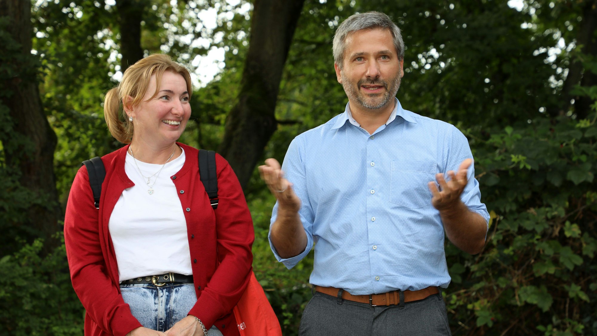 Zwei Menschen stehen vor grünen Sträuchern und Bäumen und sprechen.