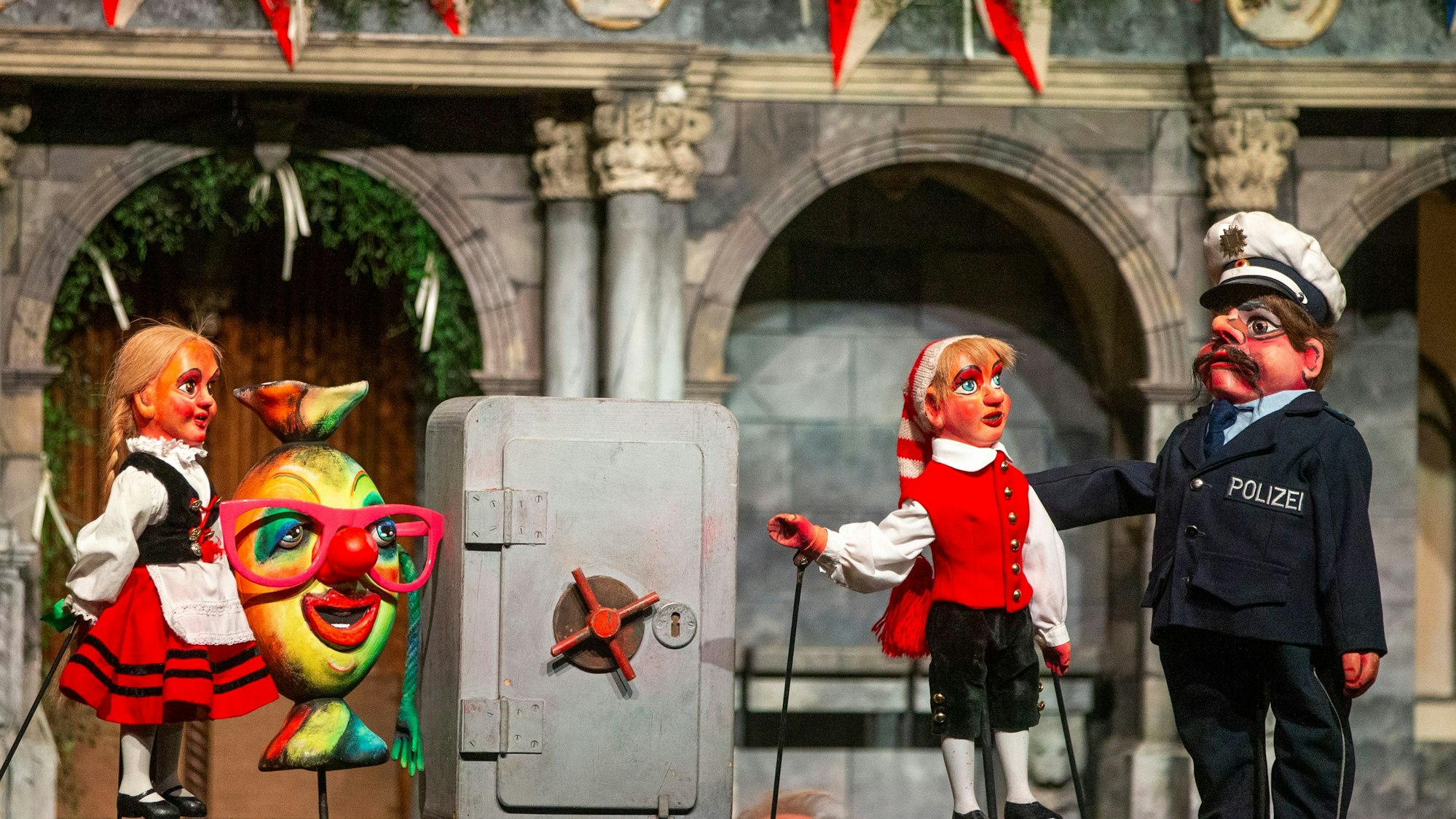 Die drei Puppen stehen auf der Bühne, dazwischen steht ein Tresor.