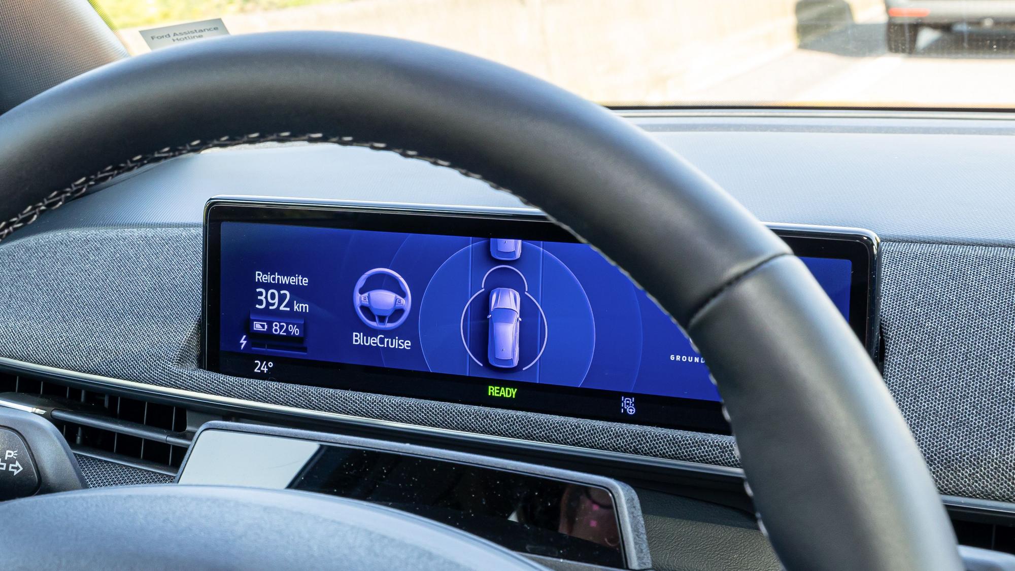 Man sieht die Sicht auf ein Lenkrad. Im Hintergrund auf dem Bildschirm steht „BlueCruise“.