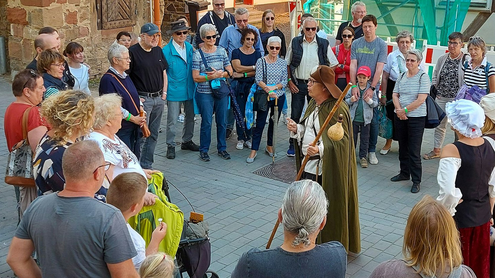 Ein Mann steht inmitten einer Ansammlung von Menschen. Er trägt die Tracht eines Jakobspilgers. Die Menschen um ihn herum tragen moderne Straßenkleidung.