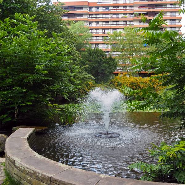 Ein Teich mit Springbrunnen in einer Parkanlage vor einem mehrstöckigen Gebäude ist zu sehen.