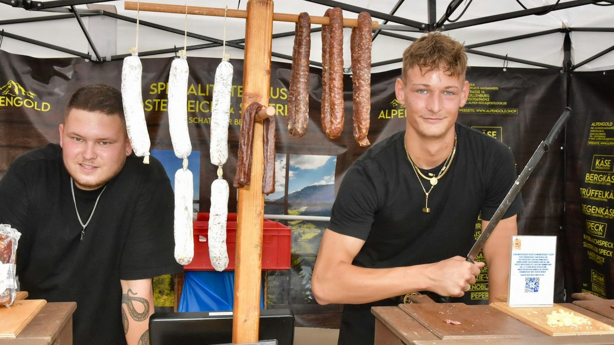 Zwei junge Männer in schwarzen T-Shirts an einem MarkTstand. An einem Holzgestell sind Dauerwürste aufgehängt. Einer der Männer hält ein großes Schinkenmesser.