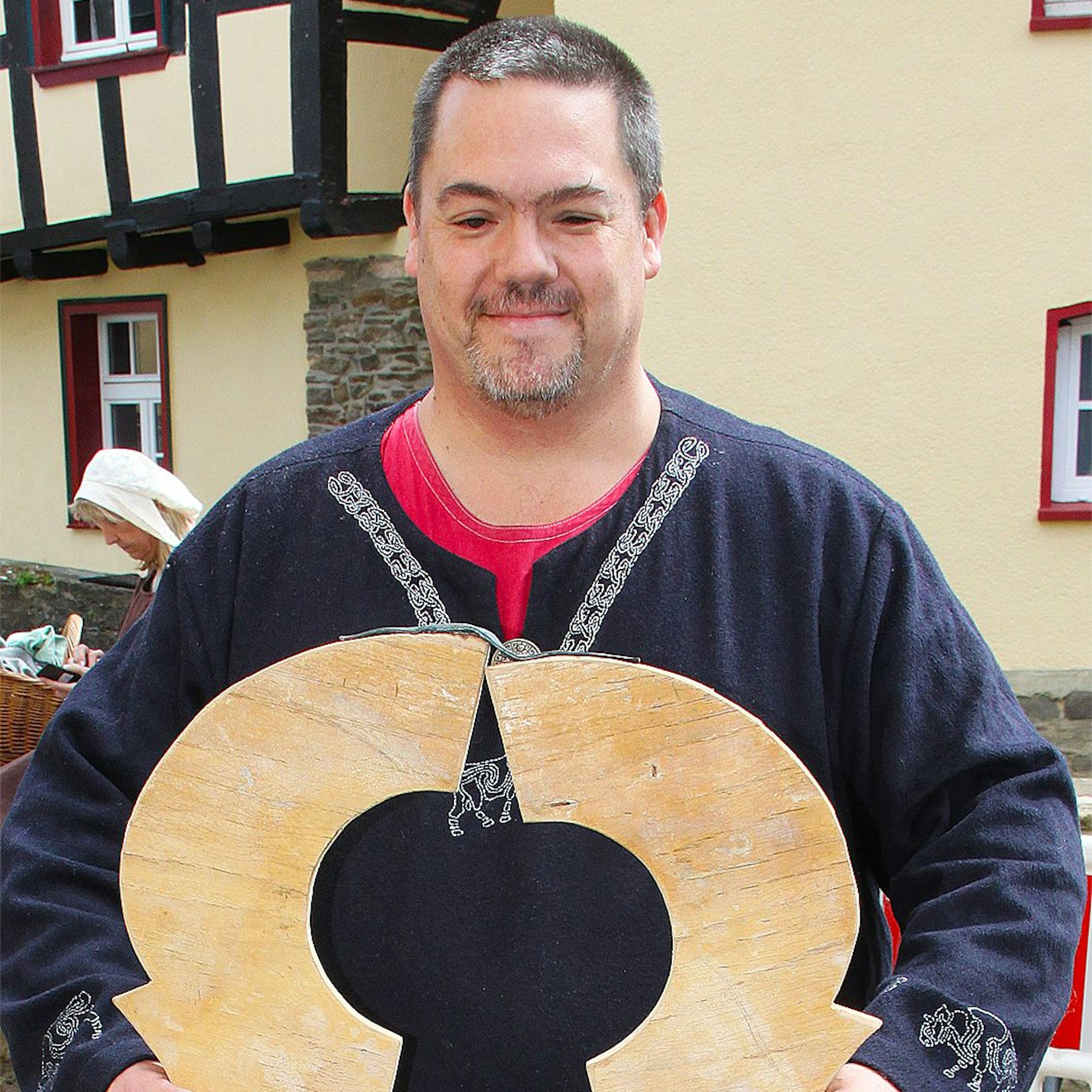 Ein Mann trägt mittelalterliche Kleidung. In seinen Händen hält er ein hölzernes Folterinstrument.