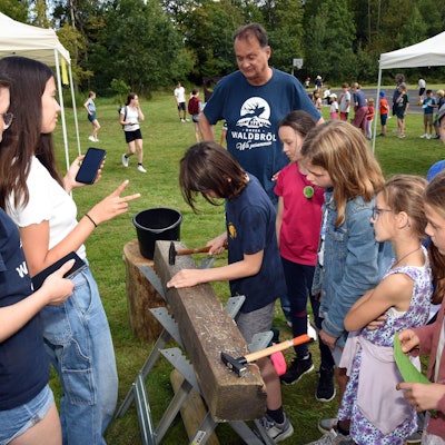 Kinder stehen um einen Nagelbalken herum, ein Kind schwingt den Hammer.