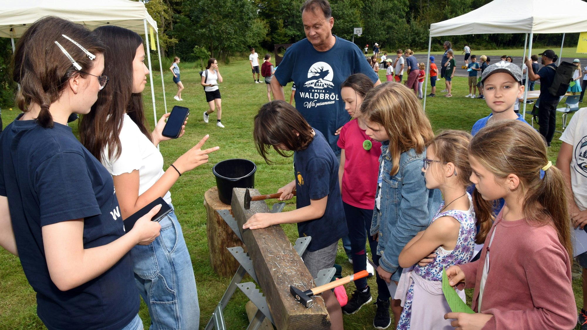 Kinder stehen um einen Nagelbalken herum, ein Kind schwingt den Hammer.