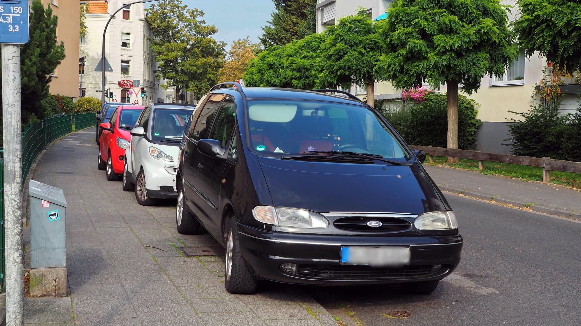 Mehrere Autos parken hintereinander an einer Straße, sie stehen halb auf dem Bürgersteig.