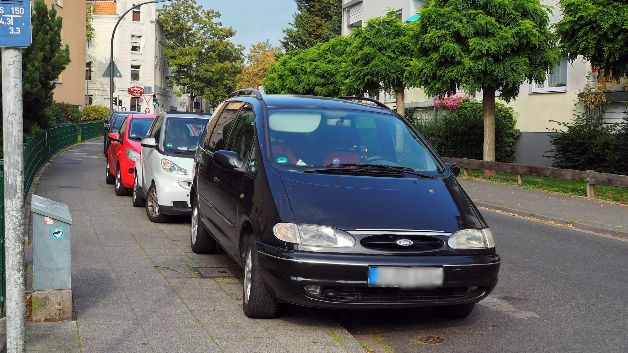 Mehrere Autos parken hintereinander an einer Straße, sie stehen halb auf dem Bürgersteig.