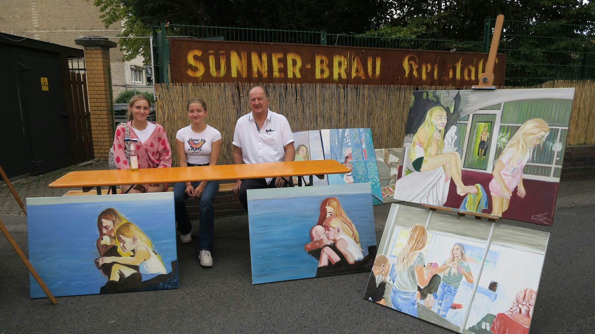 Drei Menschen sitzen an einem Stand mit Gemälden.