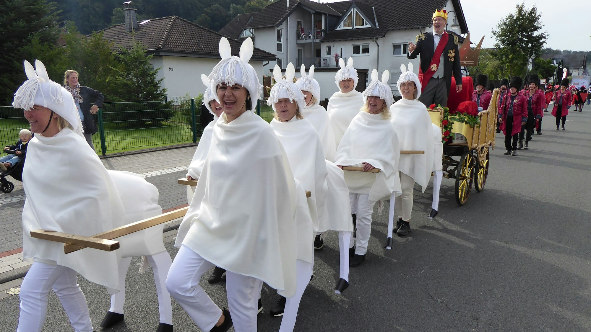 Ein Mann steht mit Krone und Schärpe auf einer Kutsche, die von Menschen in weißen Pferdekostümen gezogen wird.