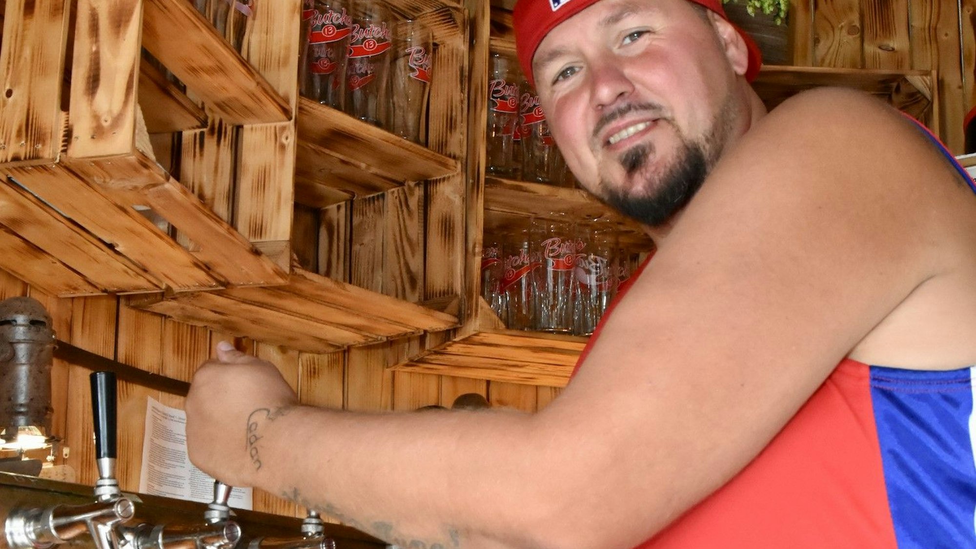 Ein junger Mann mit Bart zapft Bier. Er trägt eine umgedrehte Kappe und ein ärmelloses T-Shirt. Im Hintergrund stehen Gläser in einem Regal aus Holzkisten.