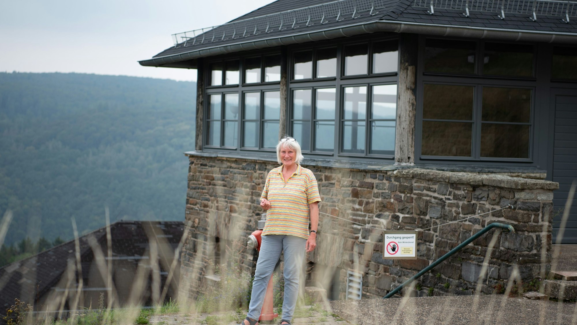 Eine Frau steht in einem gelb-gestreiften T-Shirt vor einem gemauerten Haus mit vielen Fenstern. Im Hintergrund ist ein bewaldeter Hügel zu erkennen.
