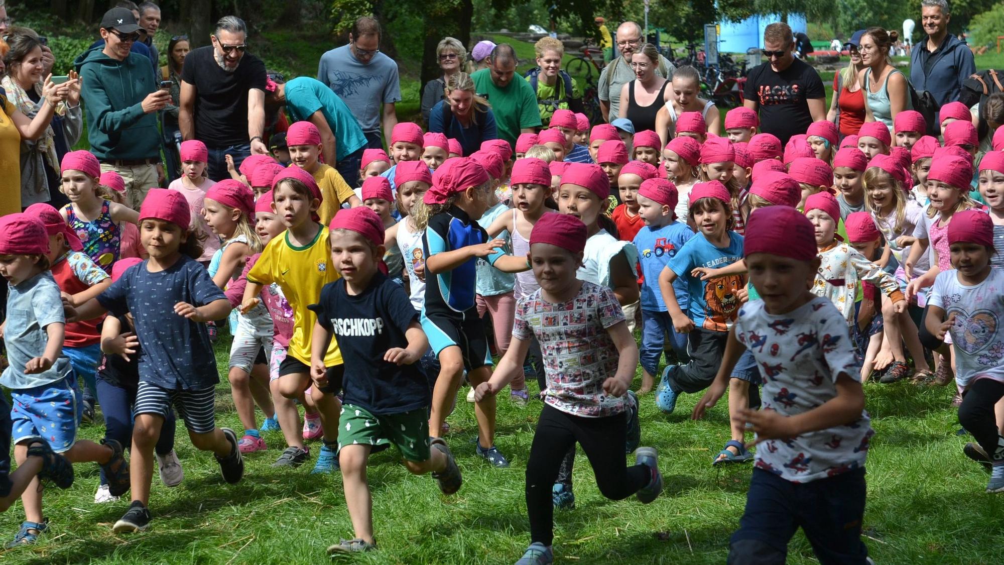 Eine große Gruppe Kinder läuft über eine Wiese. Sie alle tragen pinke Tücher um den Kopf. Im Hintergrund stehen einige Eltern.