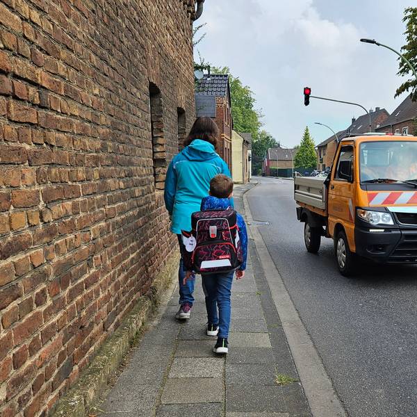Eine Mutter und ihr Sohn, der einen Ranzen trägt, gehen auf einem schmalen Gehweg neben einer Straße, auf der ein Auto an ihnen vorbeifährt.