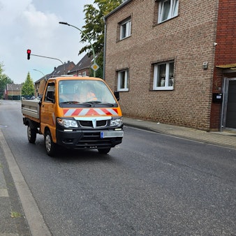 Eine Mutter und ihr Sohn, der einen Ranzen trägt, gehen auf einem schmalen Gehweg neben einer Straße, auf der ein Auto an ihnen vorbeifährt.