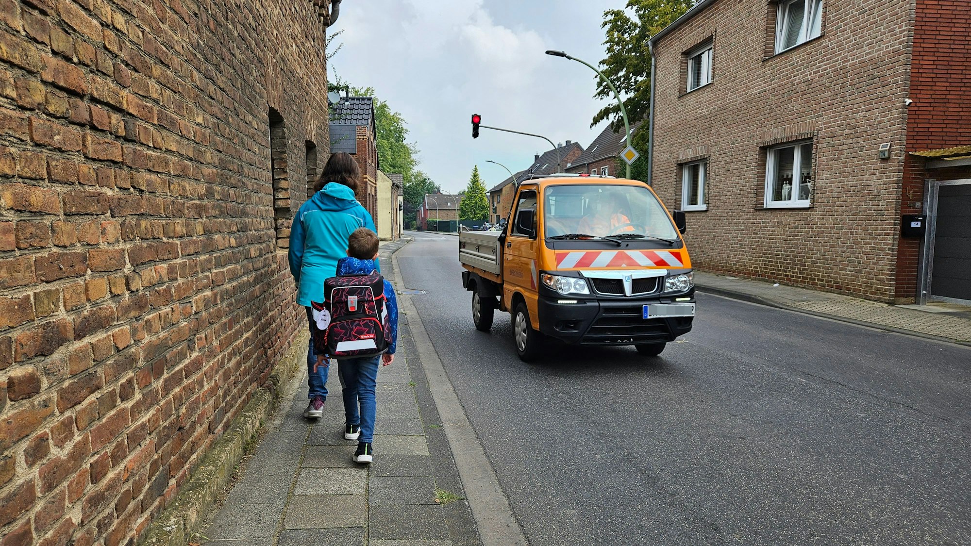 Eine Mutter und ihr Sohn, der einen Ranzen trägt, gehen auf einem schmalen Gehweg neben einer Straße, auf der ein Auto an ihnen vorbeifährt.