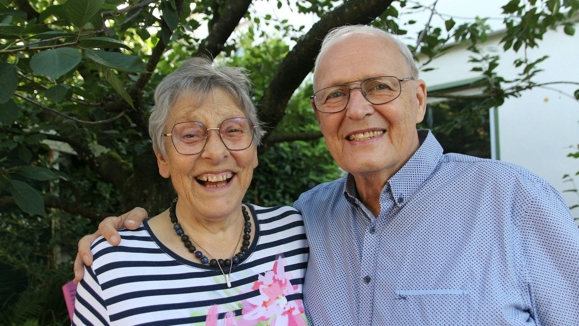 Ein Mann hat den Arm um eine Frau gelegt, sie lachen und stehen im Freien vor einem Baum.