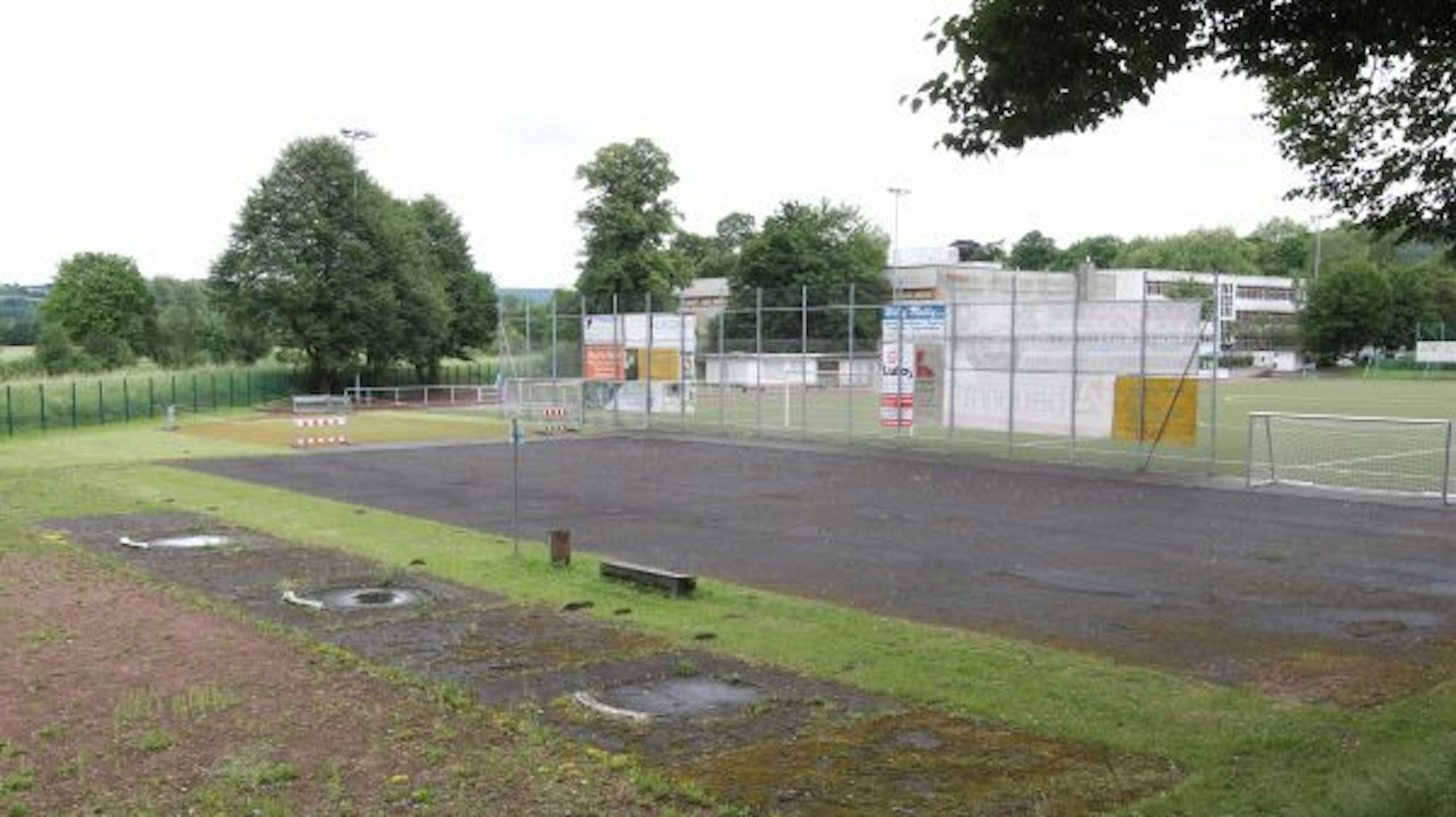Ein heruntergekommenes Kleinspielfeld unter Bäumen. Im Hintergrund ein Fußballplatz.